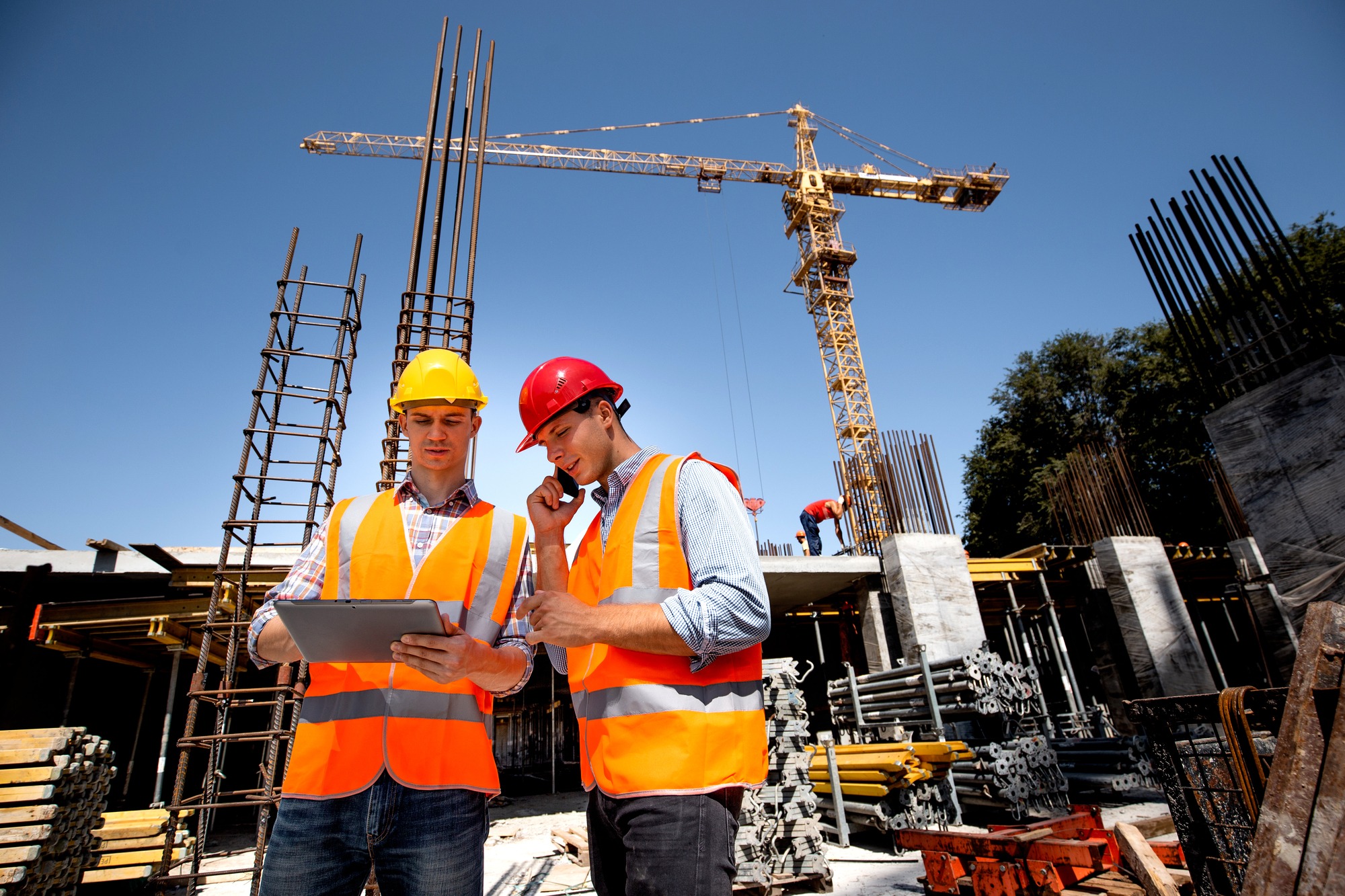 Structural engineer and architect dressed in orange work vests and hard hats discuss the construction process by the phone and use tablet on the open building site